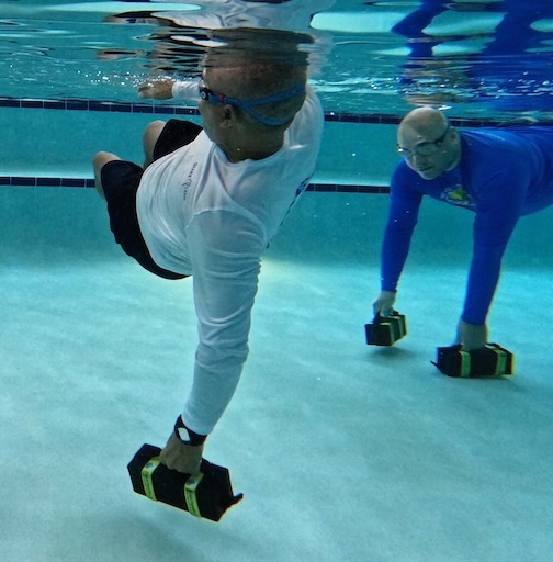 2 men under water in swimming pool holding onto rubber bricks which have taken them off balance
